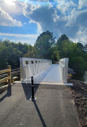 Eine Fußgängerbrücke mit Metallgeländer, einem befestigten Gehweg und einer natürlichen Landschaft mit Bäumen und einem Gewässer.