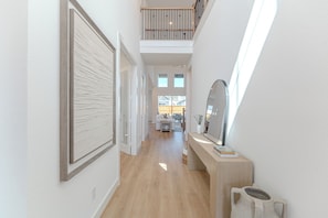 Ein Korridor mit Holzboden, einer weißen Wand mit Strukturpaneel, einem kleinen Tisch mit Spiegel und Blick in einen Wohnbereich mit Sofa und Fenster.