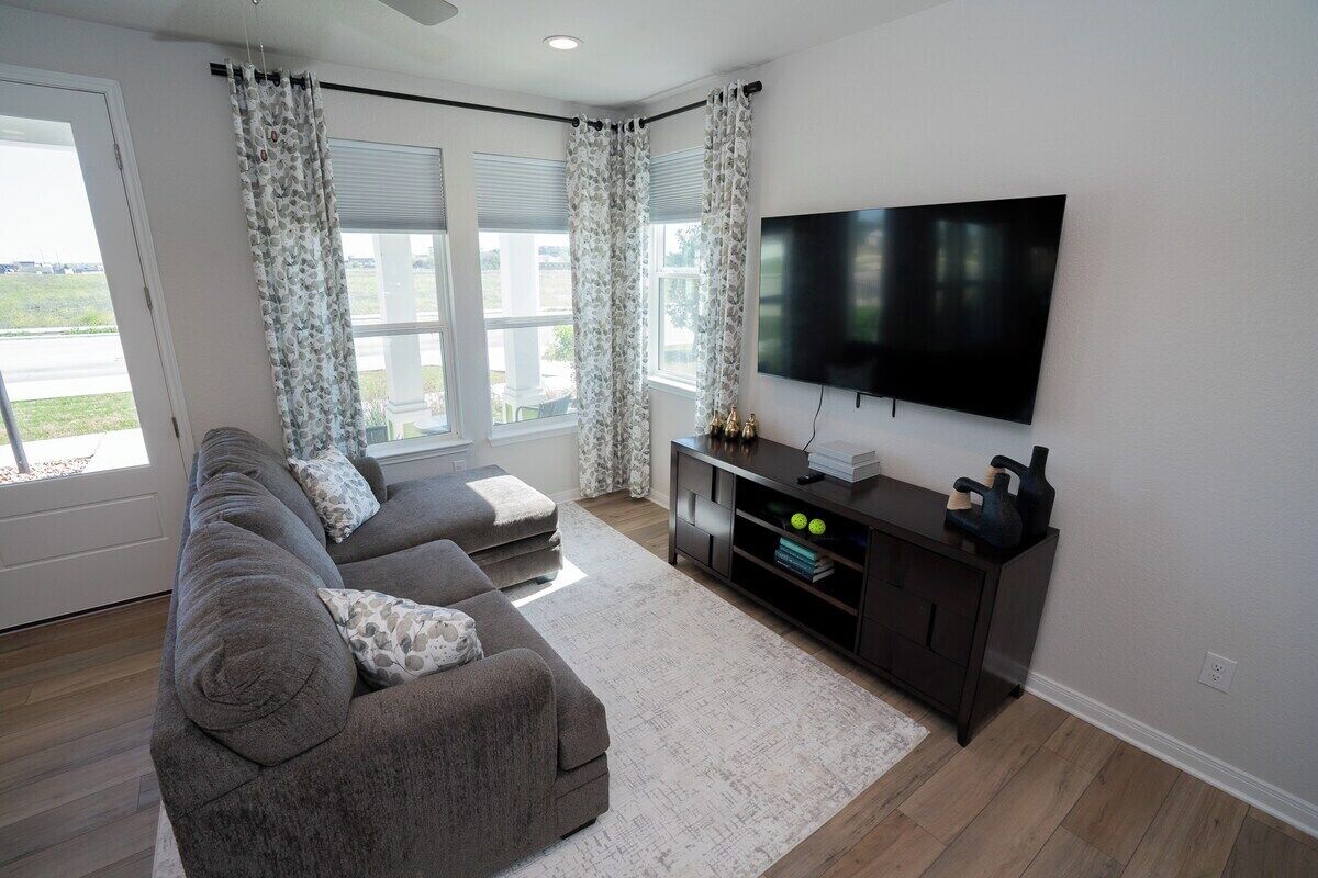 A living room with a sectional sofa, a flat-screen TV, a dark wooden entertainment unit, and a large window with curtains.