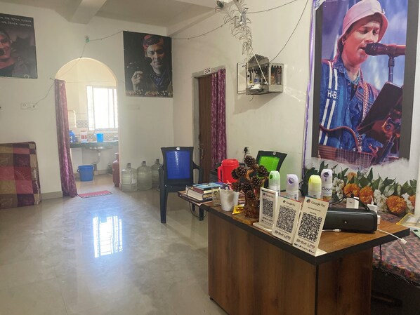 A room with a wooden reception desk, a blue chair, large water containers, a green chair, and a poster on the wall. - Maa Durga Homestay (Guwahati)