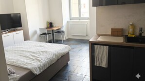 A bedroom with a bed, a desk, a chair, a TV, a window, and a kitchenette.