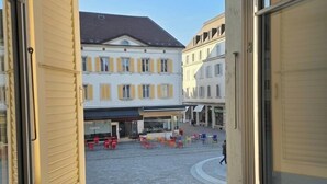 A view through a window with shutters open, revealing a courtyard with buildings, outdoor seating, and a clear sky.
