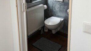 A compact bathroom with a toilet, a radiator, a window, and a wall-mounted electrical outlet.