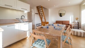 A modern kitchen with white cabinets, a wooden staircase, a dining table with blue chairs, and a living area with a brown sofa. - Renovated apartment with sea view - Trébeurden (Trébeurden)