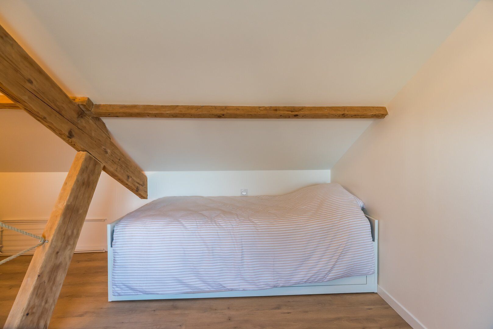 Quarto pequeno com cama de solteiro, teto inclinado e vigas de madeira aparentes.
