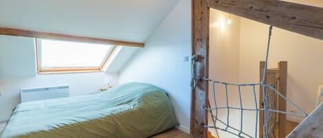 A room with a sloped ceiling, a bed, a small table with a lamp, and a rope ladder leading to a loft area.