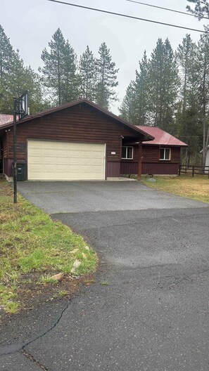 A two-story log cabin with a garage, surrounded by a gravel driveway and a grassy area. - McCall Gem, Pet Friendly,  Fully Fenced, Fire Pit! (McCall)
