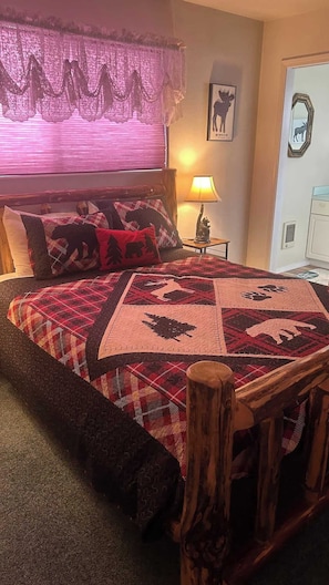 Una cama con colcha estampada de animales, cabecera de madera, lámpara de noche y ventana con cortinas de encaje.