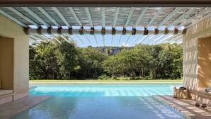 Una piscina amb una pèrgola a sobre, envoltada de vegetació exuberanta i amb una muntanya al fons.