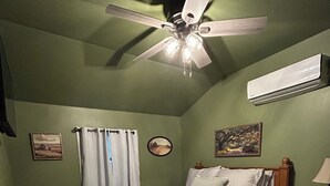 A bedroom with a bed, a ceiling fan, an air conditioner, a nightstand, a lamp, and artwork on the walls.