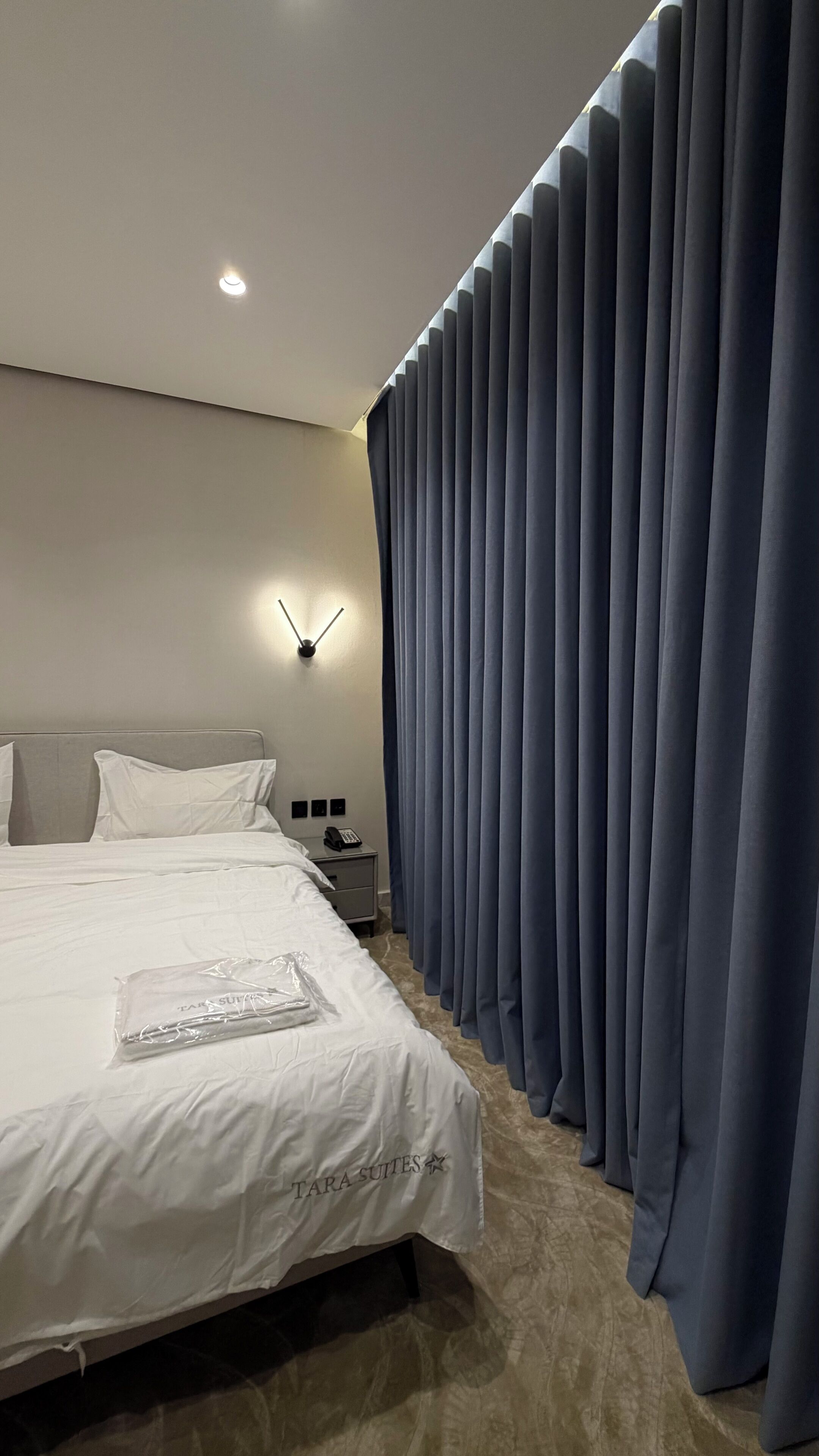 Une chambre d’hôtel moderne avec un lit soigneusement fait, une lampe fixée au mur et une grande fenêtre ornée de rideaux bleus.