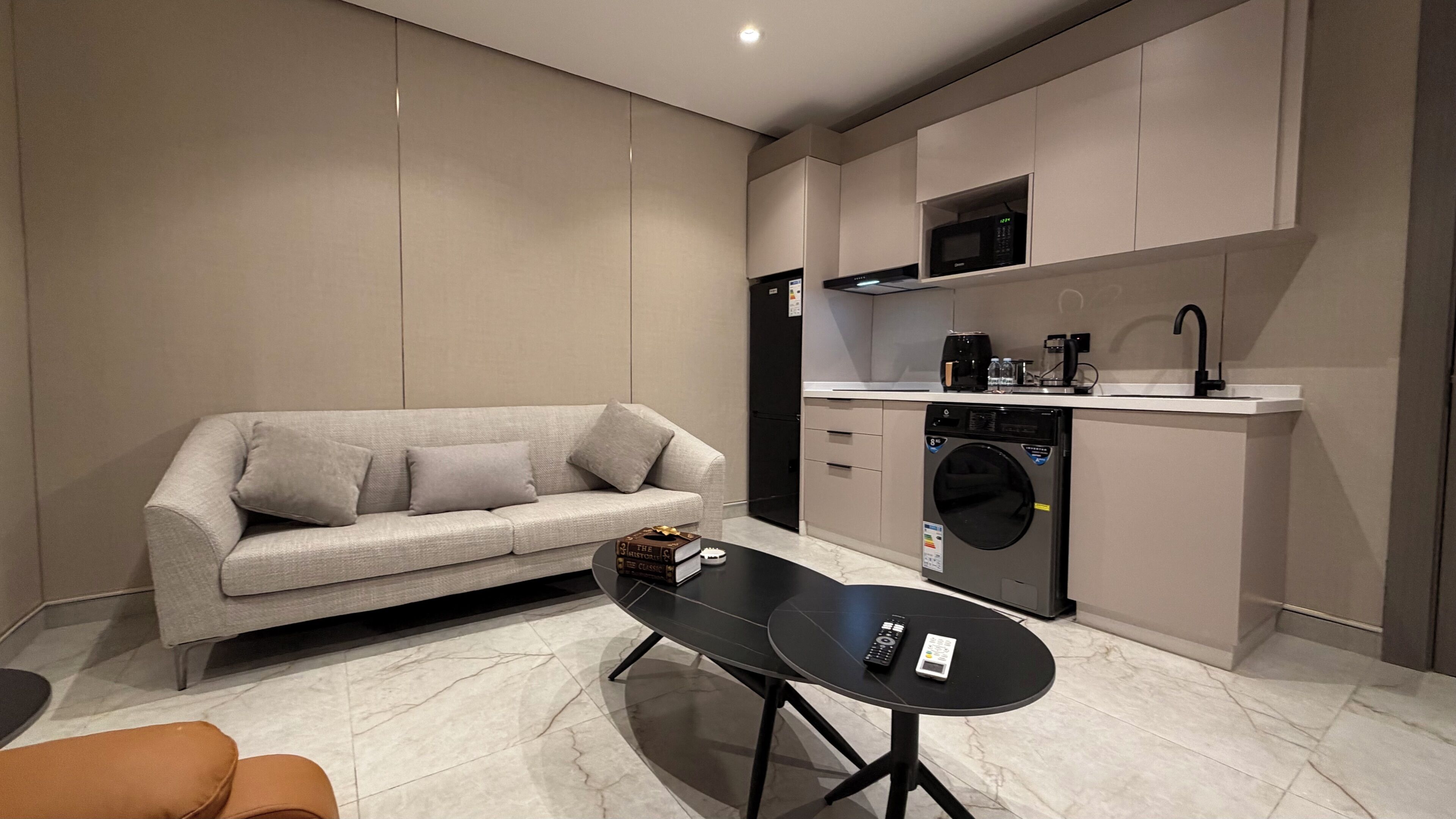 A modern living room with a sofa, a coffee table, a washing machine, and a kitchen area with cabinets and a microwave.