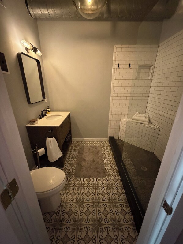 Un baño con inodoro, lavabo, espejo y ducha, todo con azulejos blancos.