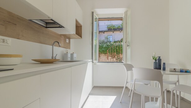 Una cocina moderna con armarios blancos, una pared decorativa de madera, un ventanal con vistas a la vegetación y una zona de comedor con sillas blancas.