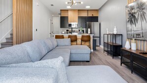 Una sala de estar moderna con un sofá modular, un comedor con taburetes de bar, una cocina con armarios de madera y un cuadro grande en la pared.