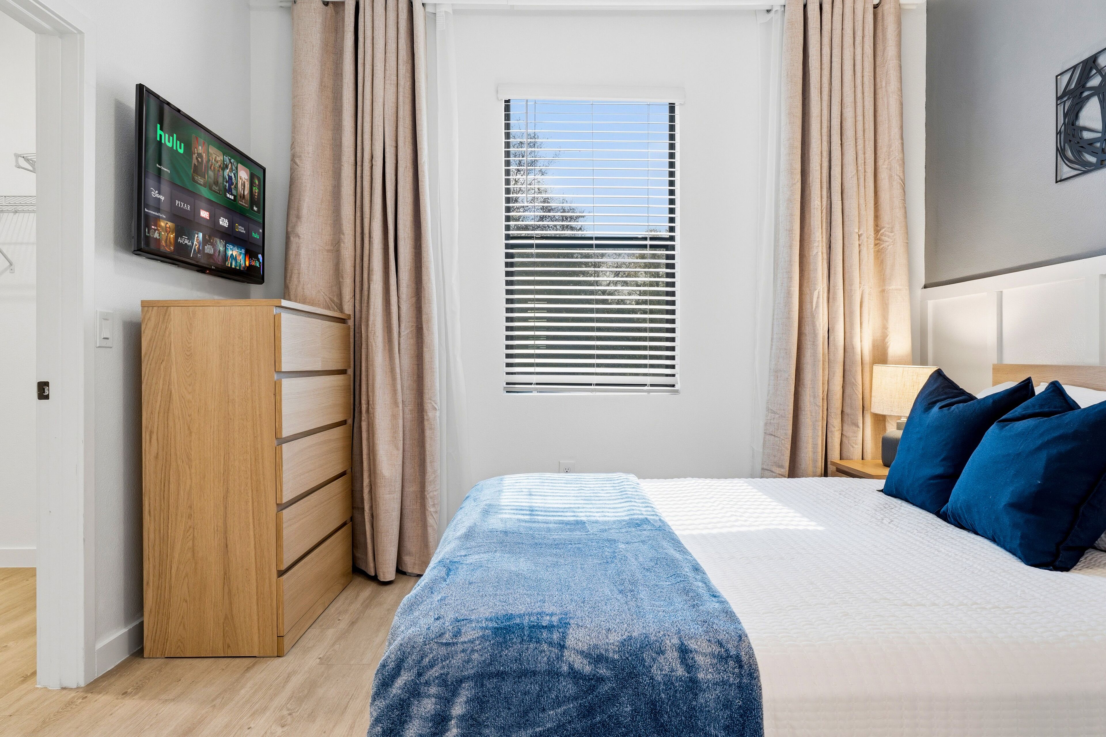 Une chambre à coucher avec un lit, un téléviseur fixé au mur, une fenêtre avec des stores et une armoire en bois.