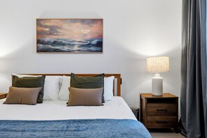 Una cama bien hecha con una manta azul, un cabecero de madera y un cuadro con una vista marina.