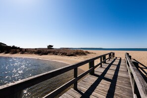 En træpromenade fører ned til en strand med klart vand og en blå himmel.