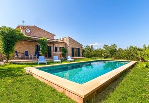 A rectangular swimming pool with a stone border, surrounded by a well-maintained lawn and a house with a stone facade and green shutters. - Ses Veles by Rentallorca (Binissalem)