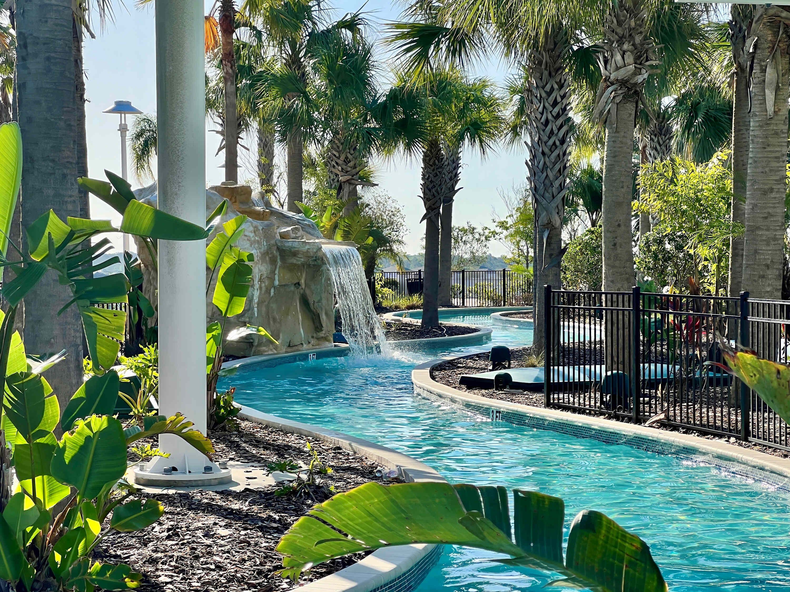 A swimming pool with a waterfall feature, surrounded by palm trees and a fence.