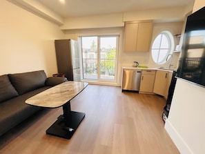 A modern living space with a sofa, a dining table, a kitchen area with a dishwasher, and a large window providing a view of the outside. - Modern Spacious 2 Bed 2 Bath Townhouse (Vancouver)