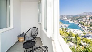 Un balcón con dos sillas y una mesita pequeña, con vistas a un pueblo costero que incluye un puerto deportivo y edificios.