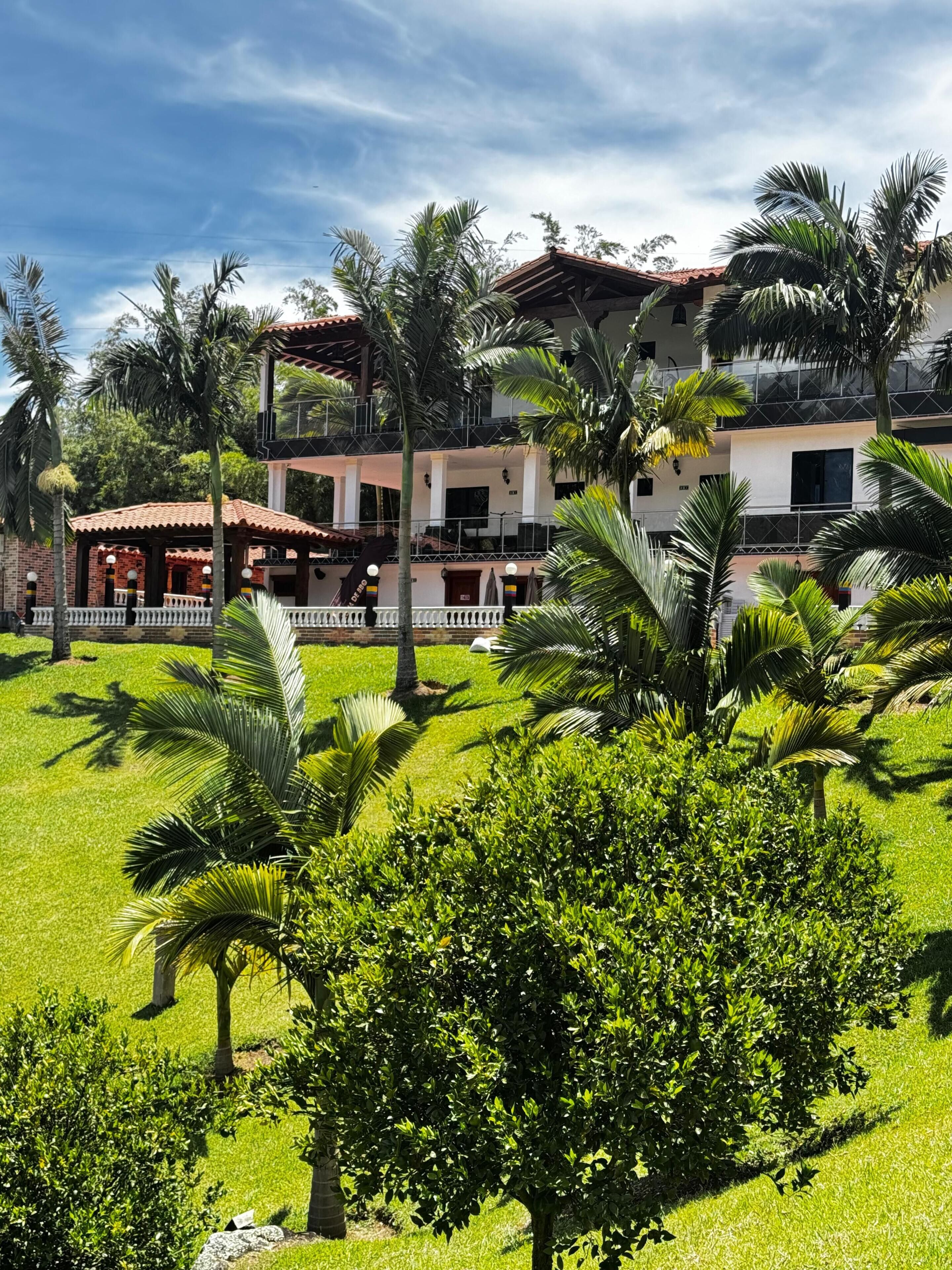 Un hotel de dos plantas, con una zona de estar al aire libre cubierta y un césped cuidado con palmeras.