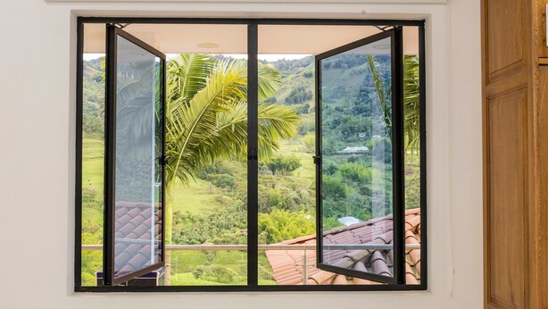A room with a large window offering a view of a lush green landscape and a palm tree. - Ecohotel Dubai Del Oriente (El Penol)