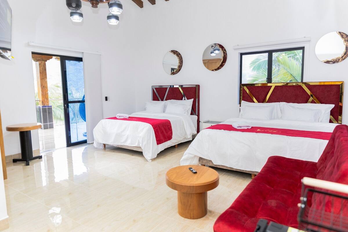 Een hotelkamer met twee bedden, een rode bank, een kleine houten tafel en een groot raam met uitzicht op palmbomen.