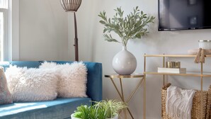 Ein Wohnzimmer mit einem blauen Sofa, einem runden Couchtisch mit einer Pflanzenbowl, einem an der Wand montierten Fernseher und einer Stehlampe.