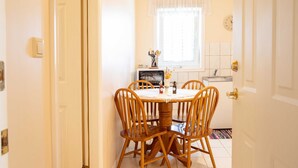 Un área de comedor con una mesa redonda de madera, seis sillas, una ventana con cortina blanca y una cocina visible al fondo.