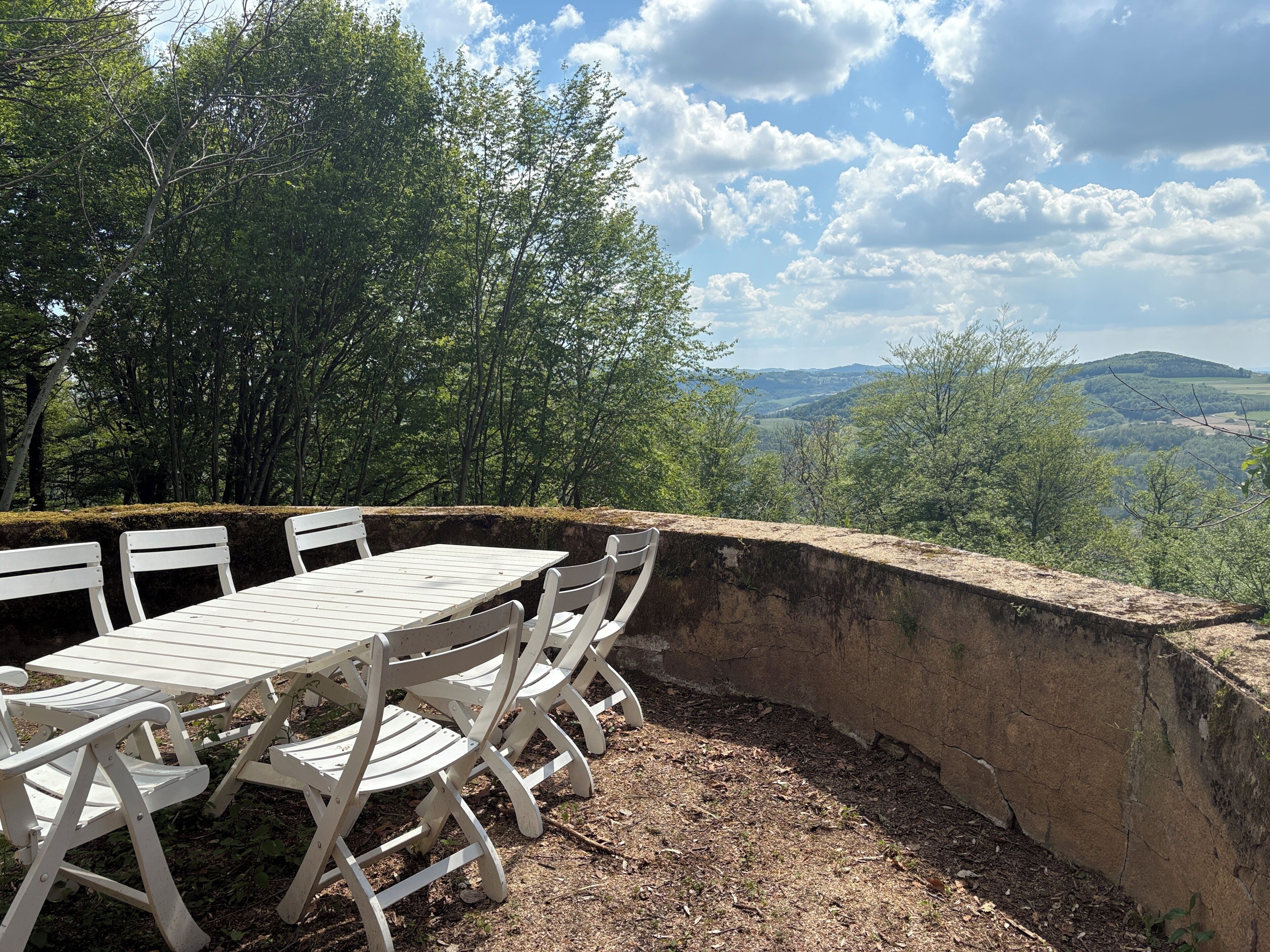 A picnic table with chairs, surrounded by a stone wall, overlooking a forested valley with distant hills.
