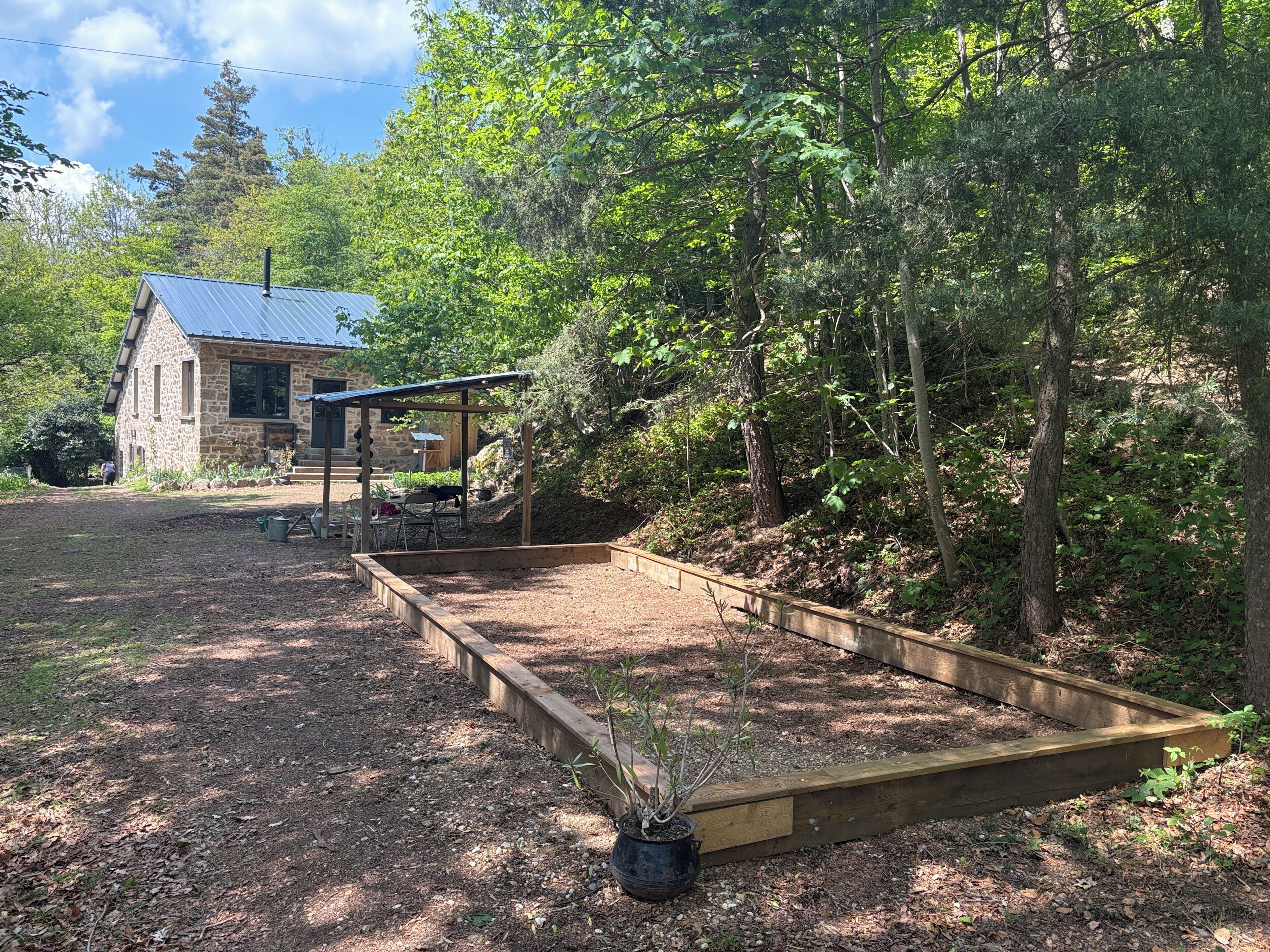 Una casa de piedra con techo de metal, un muelle de madera y un jardín con un cantero elevado y una planta en maceta.