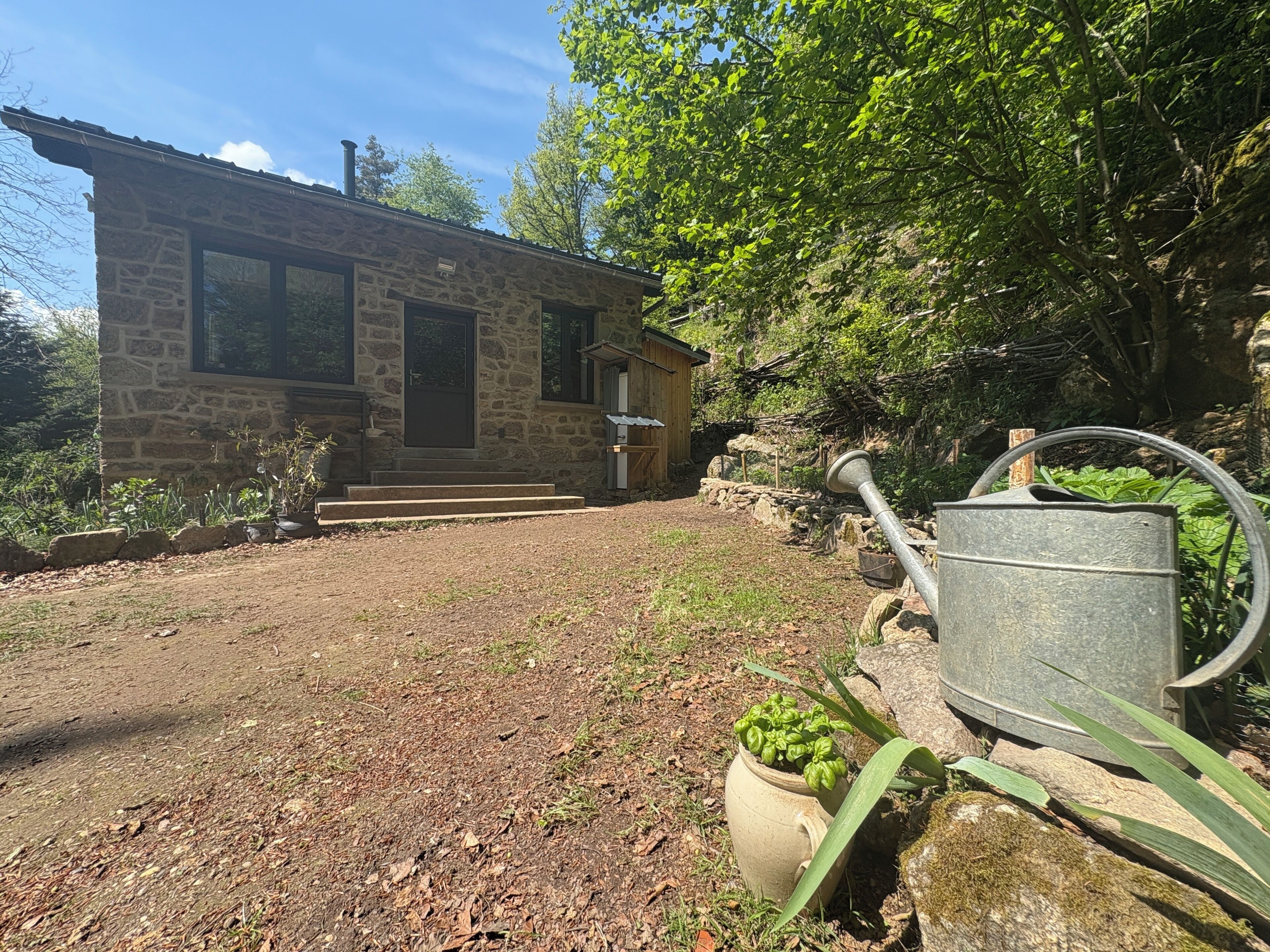 Une maison en pierre avec une porte d’entrée, des marches menant à celle-ci et un jardin agrémenté d’un arrosoir et de plantes en pot.