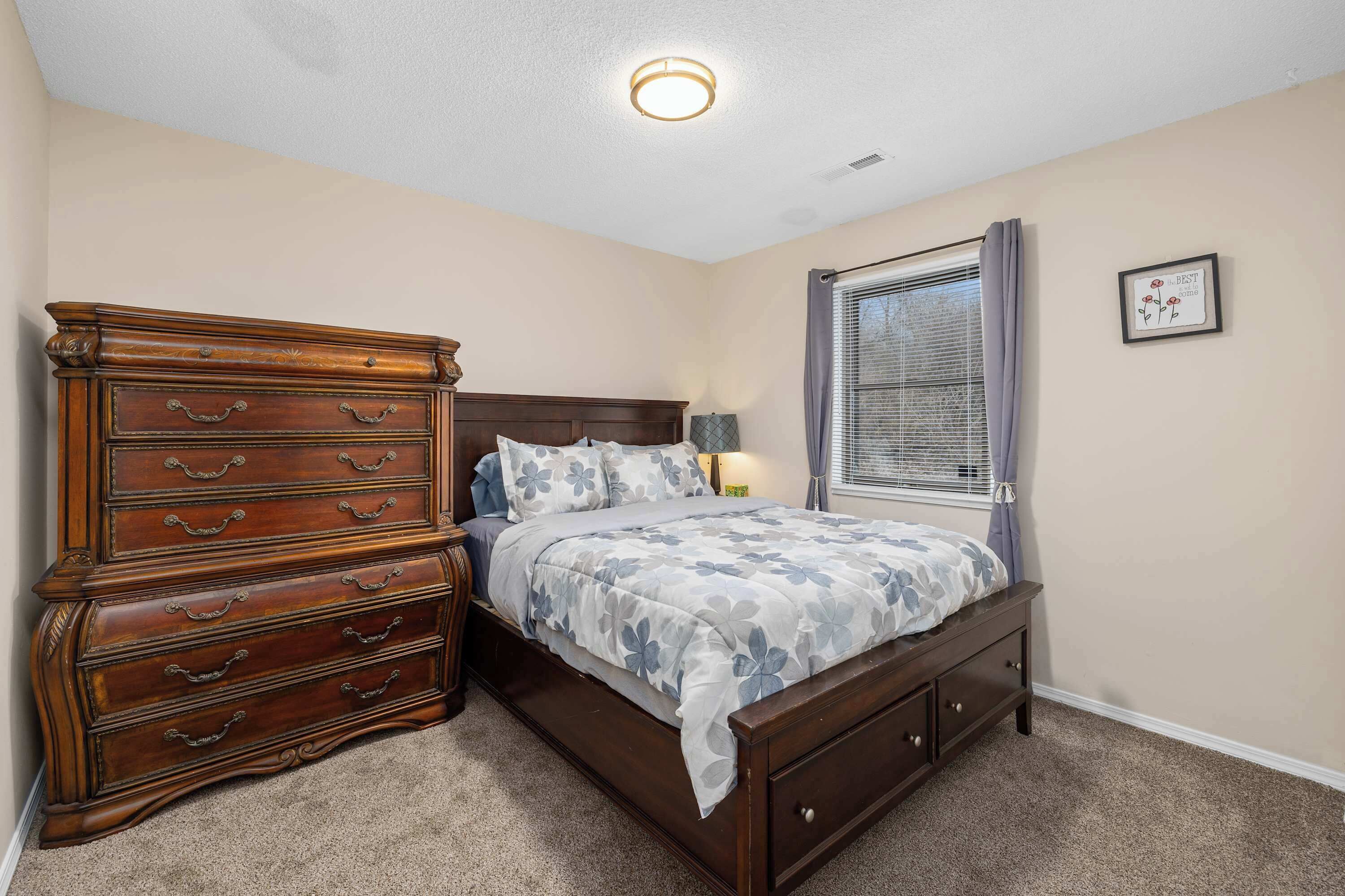 A bedroom with a wooden dresser, a bed with floral bedding, a nightstand, a lamp, and a window with curtains.