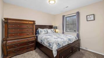 A bedroom with a wooden dresser, a bed with floral bedding, a nightstand, a lamp, and a window with curtains.