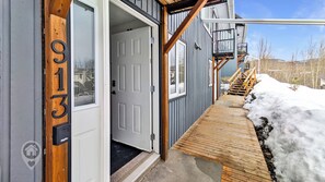 Une porte d’entrée en bois avec une plaque portant un numéro, un escalier extérieur abrité et un sol recouvert de neige.