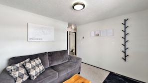 Un salon moderne avec un canapé gris, un coussin décoratif à motifs, une table basse en bois, un luminaire suspendu au plafond et une œuvre d’art accrochée au mur.