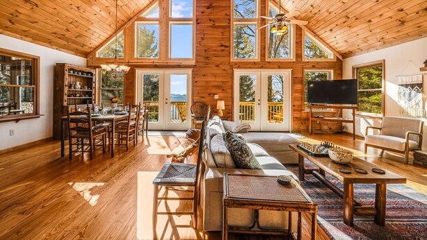 Ein geräumiger Loft-Raum mit hoher Decke, Holzbalken, großen Fenstern, einem Kamin, einer Couch, einem Esstisch und einem Fernseher.