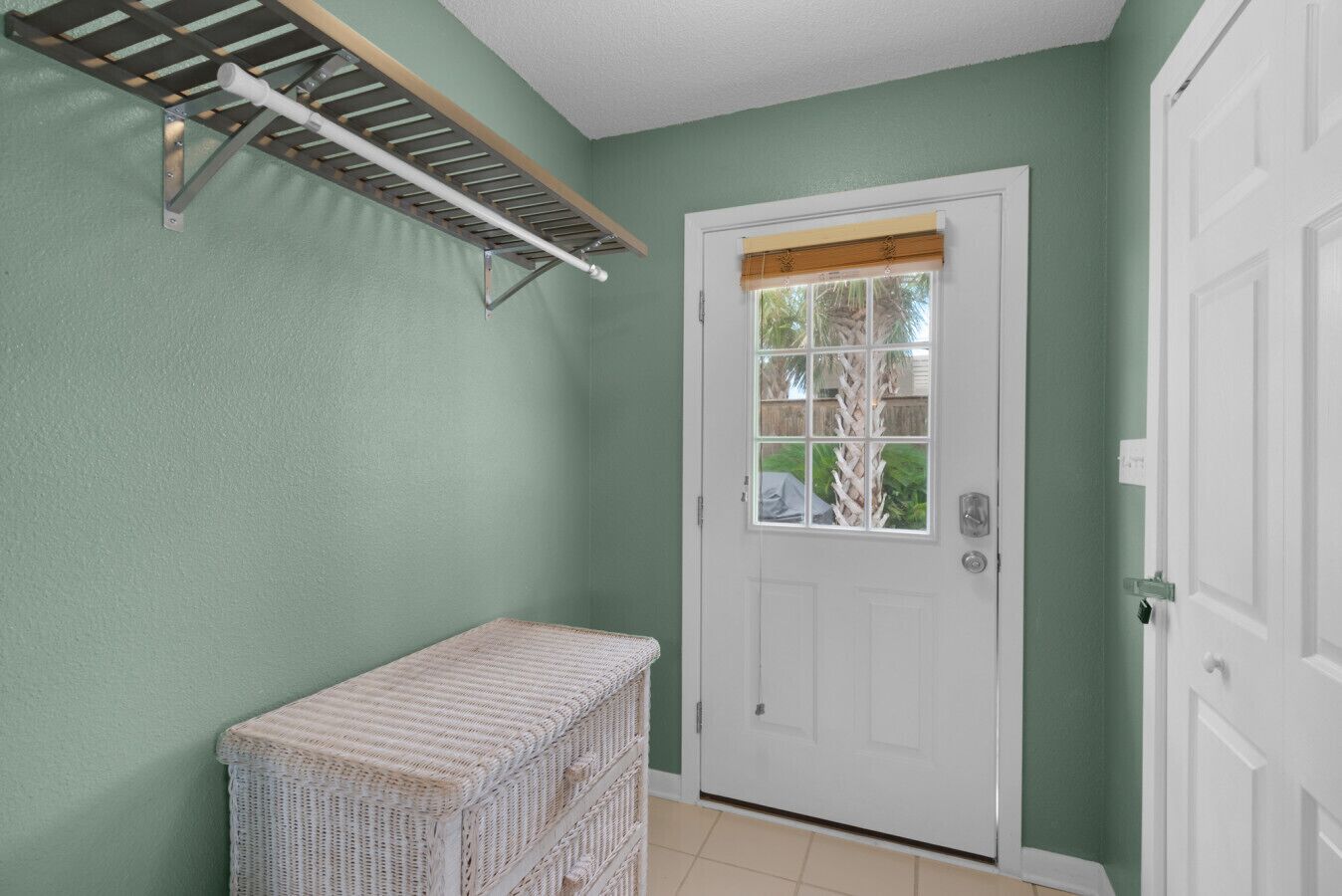 A room with a white door, a window with a view of greenery, a white wicker bench, and a drying rack on the wall.