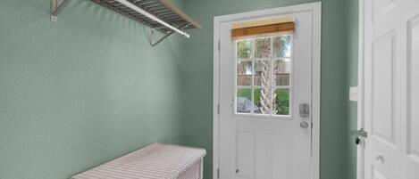 A room with a white door, a window with a view of greenery, a white wicker bench, and a drying rack on the wall.