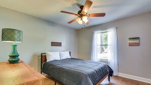 Ein Schlafzimmer mit einem Bett, Deckenventilator, Nachttisch, einem Fenster mit Vorhängen und einem Wandbild.