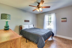 Un dormitorio con cama, ventilador de techo, mesita de noche, ventana con cortinas e imagen colgada en la pared.