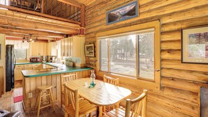 Ein Blockhauskaminareal mit einem Holzesstisch, Stühlen, einer Küchenzeile und einem großen Fenster.