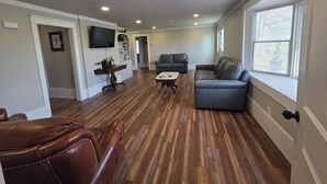 Un salon spacieux doté d’un parquet, d’un canapé gris, d’un fauteuil en cuir marron, d’un téléviseur fixé au mur et d’une fenêtre avec des stores.