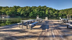 Un muelle de madera con mesas de picnic y bancos que da a un lago tranquilo, rodeado de un bosque denso.