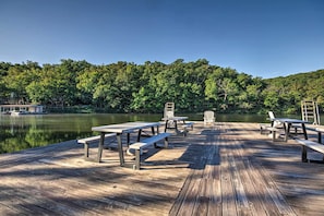 Un muelle de madera con mesas de picnic y bancos que da a un lago tranquilo, rodeado de un bosque denso.