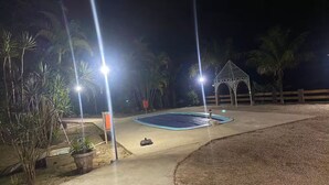 Espacio al aire libre de noche con piscina, palmeras y un pabellón.