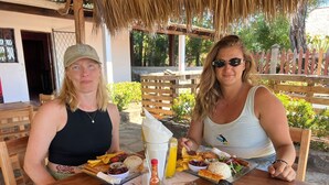 Two people seated at an outdoor table with food and drinks, a thatched roof, and a wooden fence in the background. - Sabores de mi Tierra (Popoyo)