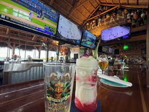 Angolo bar con diversi schermi che trasmettono eventi sportivi, un cocktail dal design tropicale e un soffitto in legno rustico.